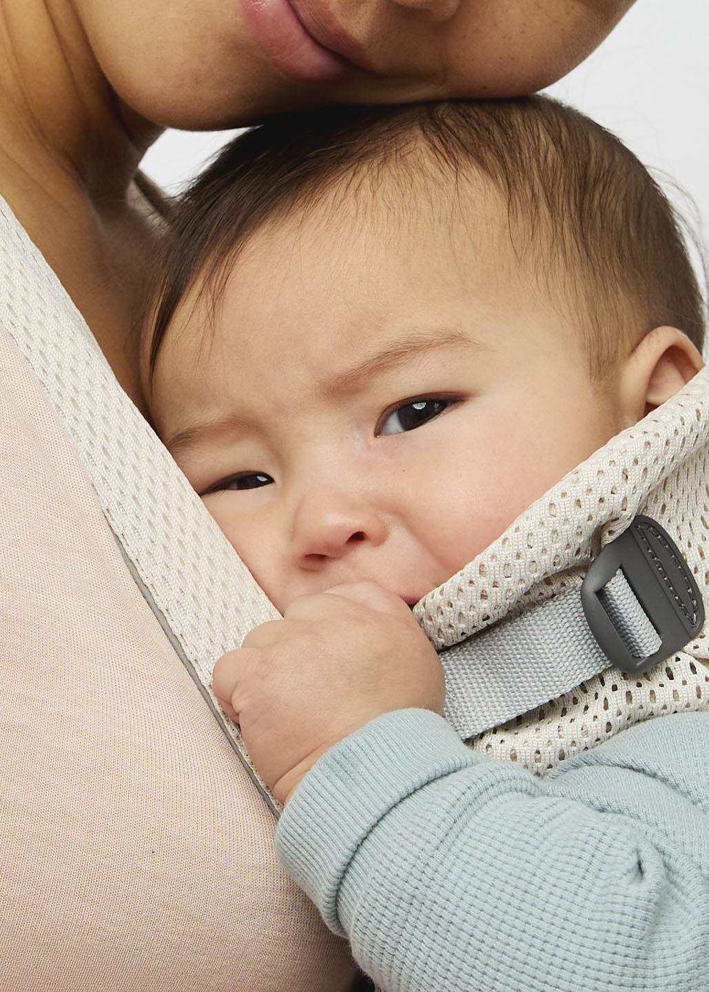 Baby cuddled against a person's chest, held securely in a white carrier, wearing a light blue shirt and looking calm.