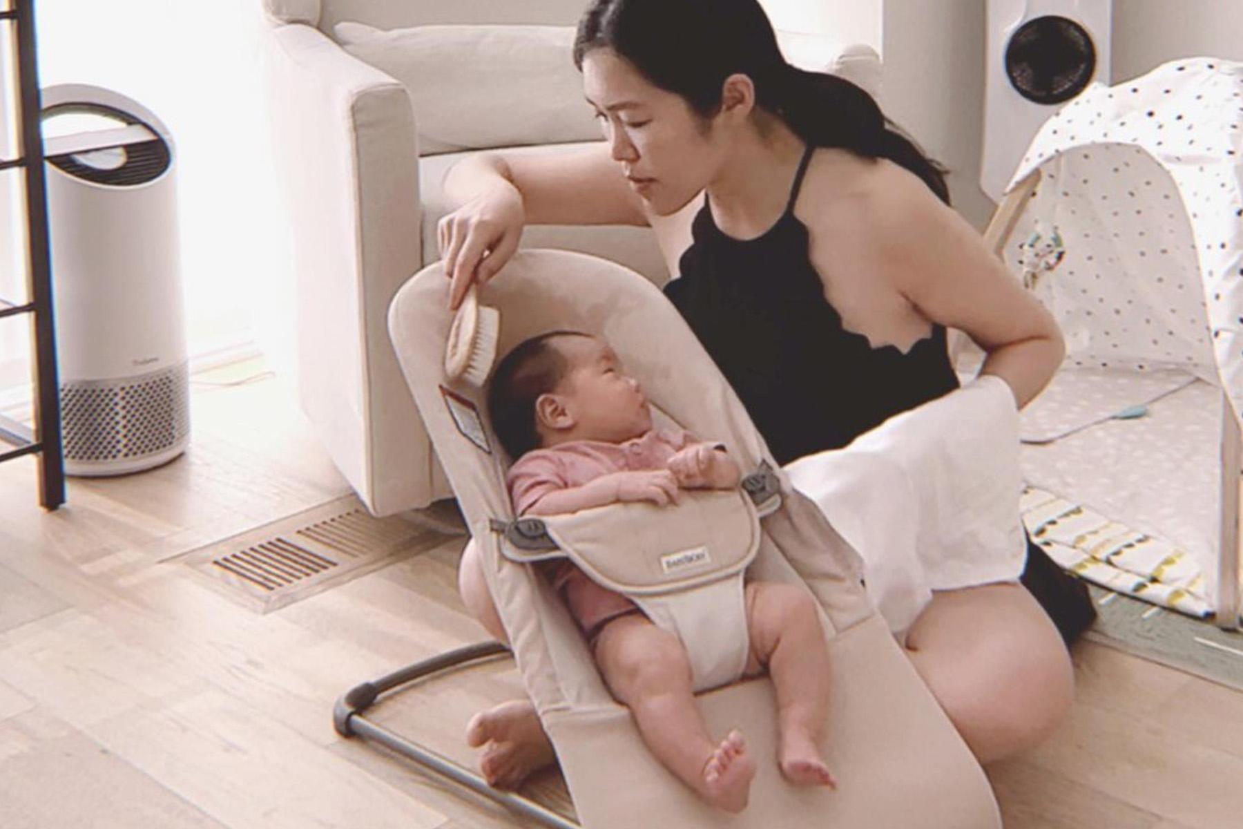 A woman sitting on the floor gently strokes a baby in a bouncer inside a cozy living room.