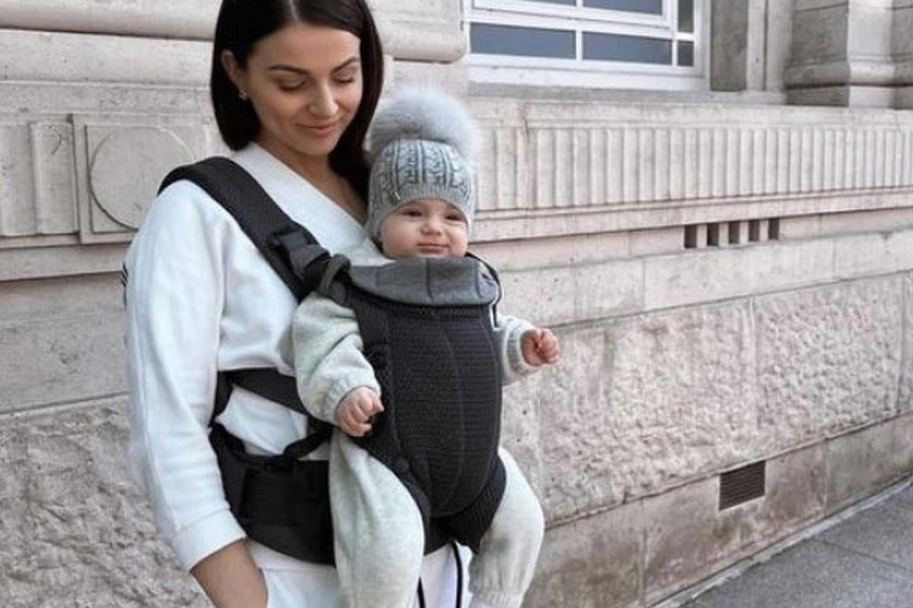 A woman stands against a stone wall wearing a black baby carrier, holding a baby dressed in a gray outfit and a knitted hat with a pom-pom.