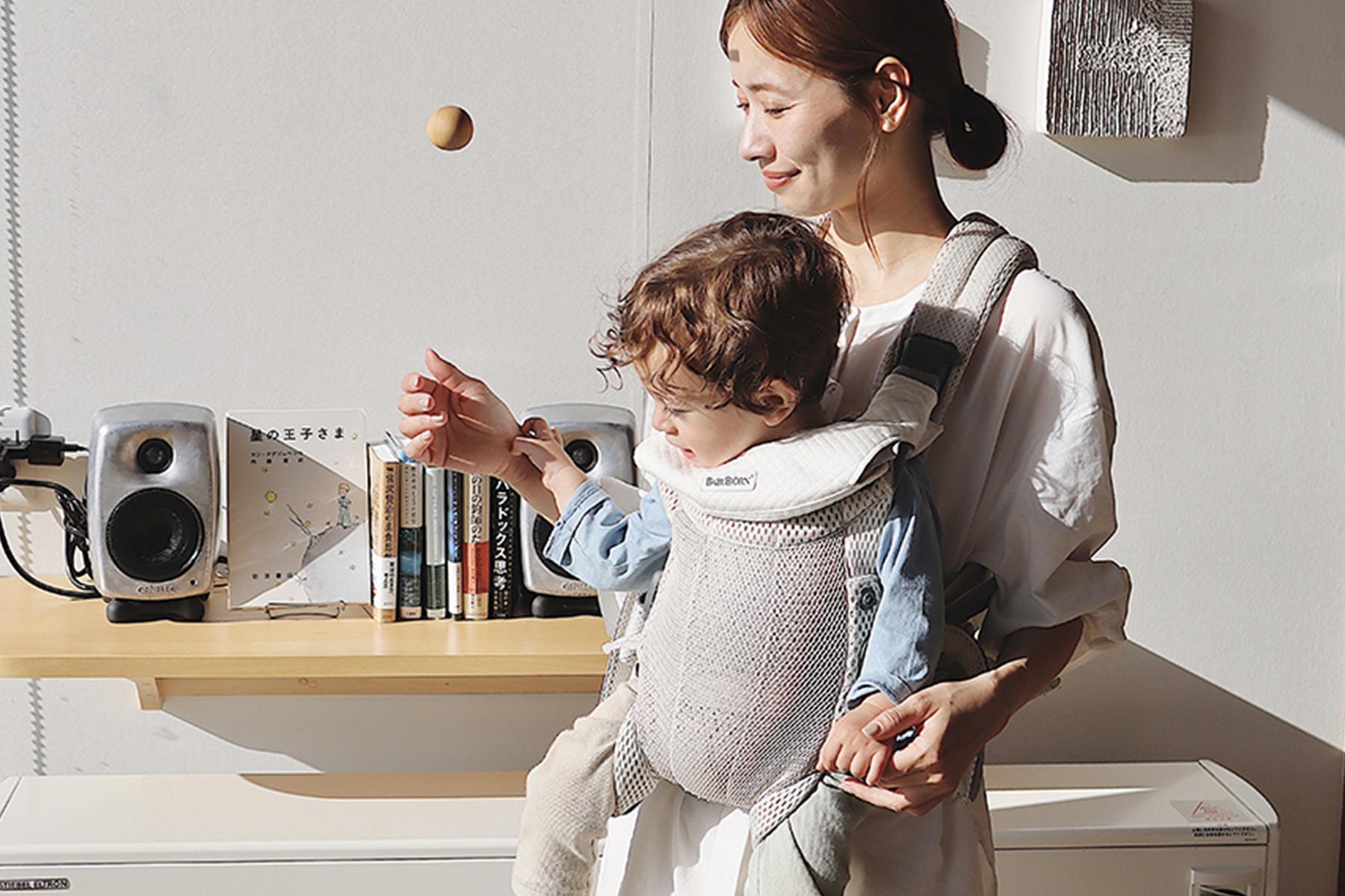 A woman smiles while holding a child in a baby carrier. They are indoors near a shelf with books and speakers, with sunlight streaming in.