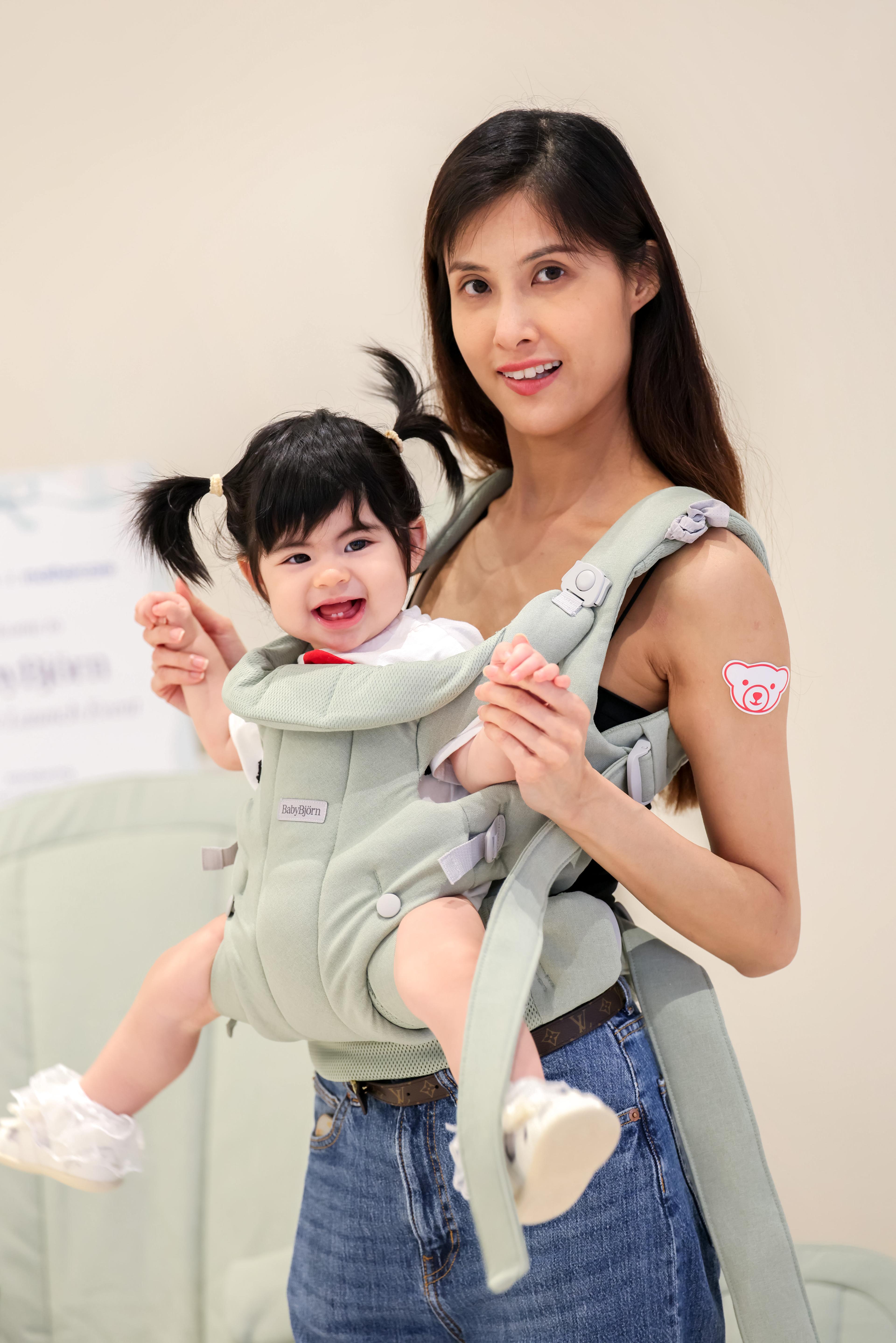 A woman smiling while carrying a laughing baby in a green baby carrier. The baby has pigtails, and the woman has a sticker on her arm.