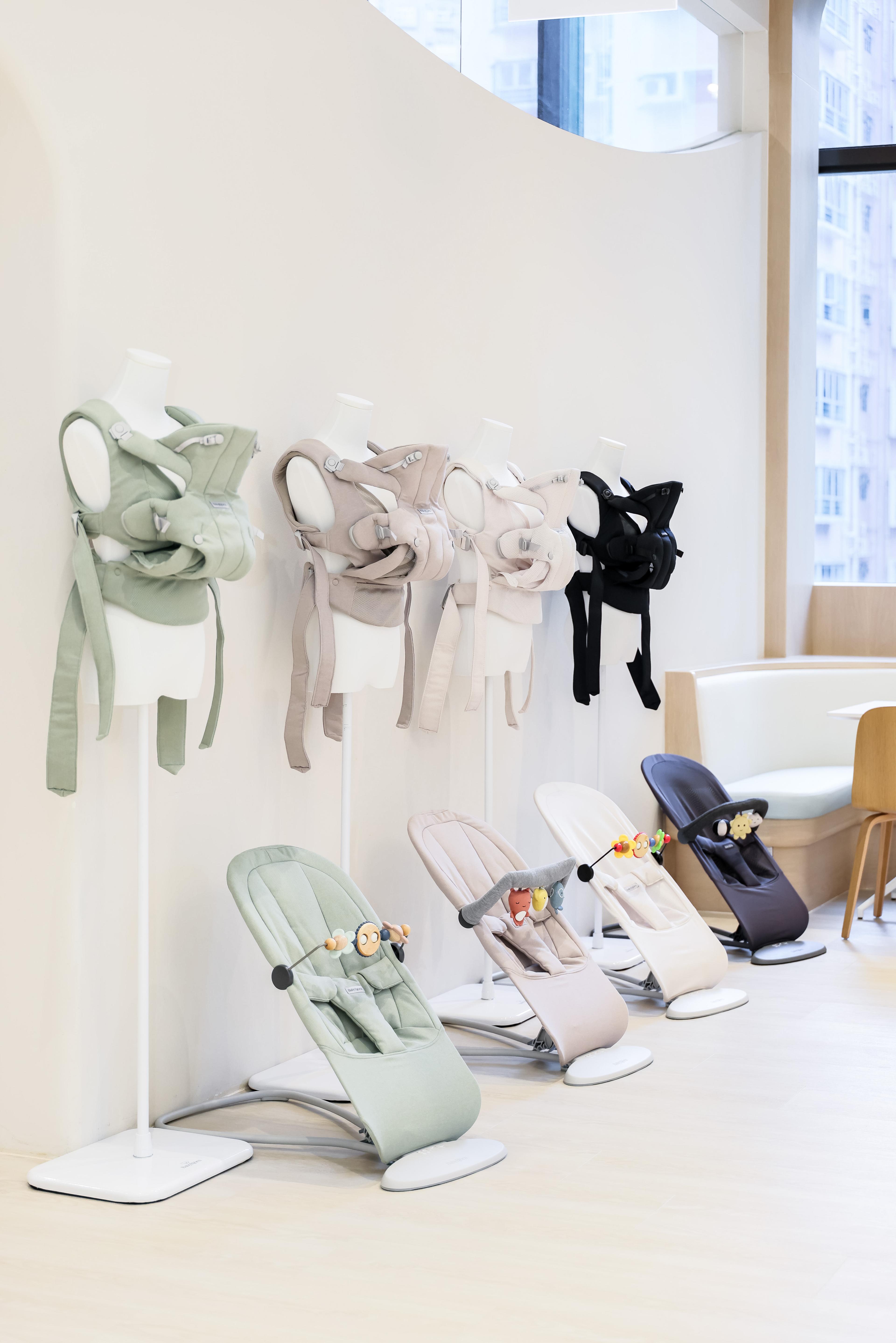 Display of pastel-toned baby carriers on mannequins and baby bouncers with toys in a modern showroom.