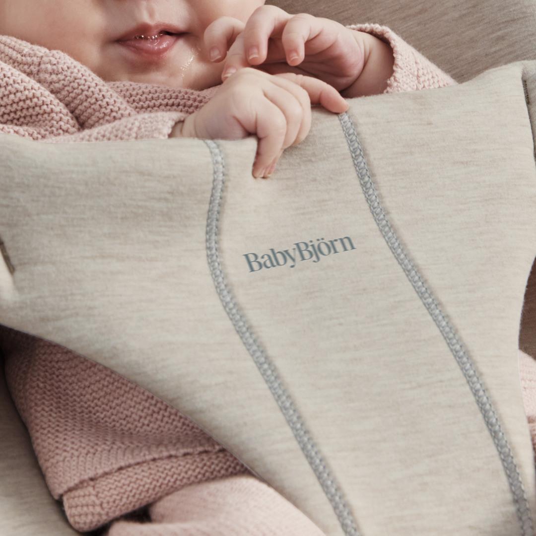 A baby wearing a pink knitted outfit is partially visible, resting in a beige BabyBjörn carrier, with fingers near the mouth.
