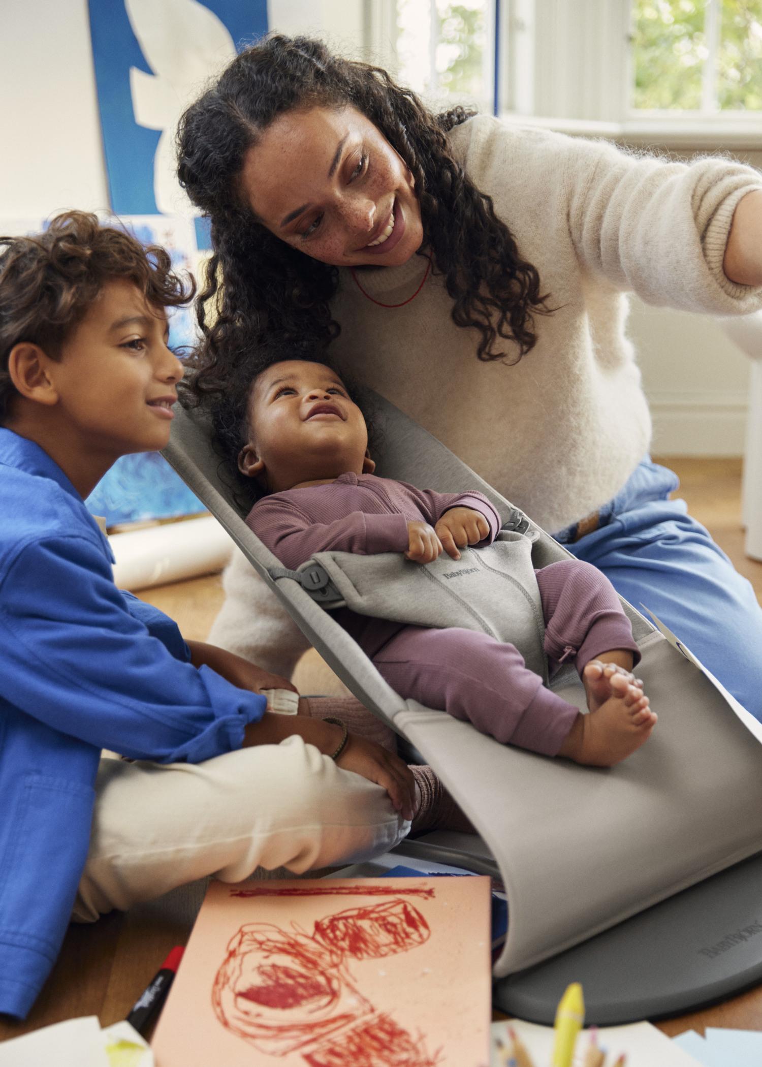 A woman and a young boy smile at a baby in a bouncer. Art supplies and a drawing are on the floor in a bright room.